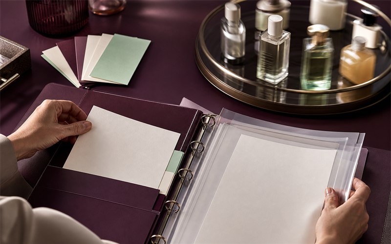 Two realistic hands moving a revised fragrance certificate into an organized compliance file beside sealed product samples and a date-stamped change-control card without readable text