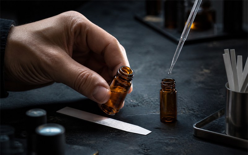 A perfumer's hand moves a small unbranded amber sample vial beside a fragrance blotter while a second vial is being gently rotated, showing controlled formula adjustment and batch review